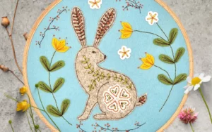 A brown hare embroidered onto a light blue felt background and placed in an embroidery hoop.